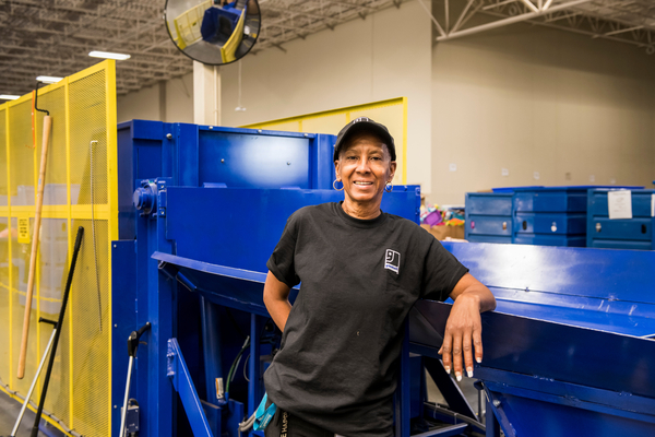 A Goodwill employee stands confidently beside a blue salvage processing machine, showcasing Goodwill's salvage and broker services.