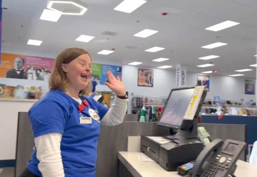 Page stands behind the Goodwill checkout counter, smiling and waving while wearing her blue Goodwill shirt and name tag. The store sales floor is visible behind her as she greets customers with her friendly, positive energy.